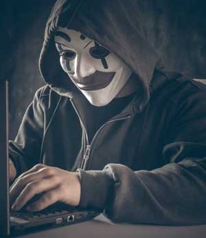 A hooded man with an “Anonymous” mask sits before a laptop computer.